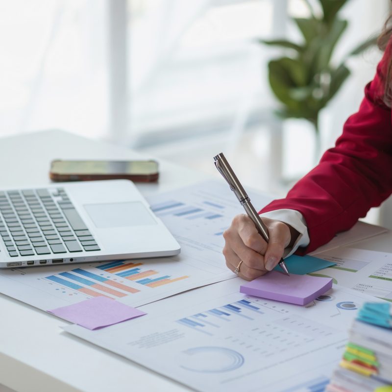 Asian businesswoman in red suit calculating company sales with calculator, laptop, and tablet on table Interior of businesswoman office at the modern workstation.